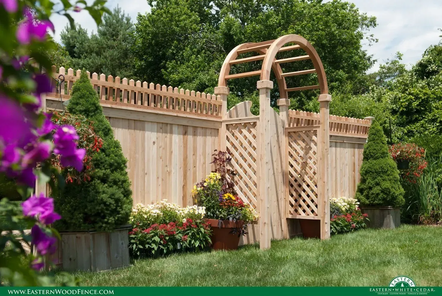 Wooden garden fence with an arched gate, surrounded by green grass, flowers, and trees.