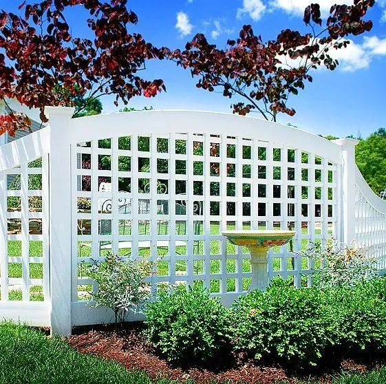 White lattice fence in a garden with bushes, birdbath, and red-leafed tree against a blue sky.