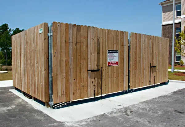 Wooden fence enclosure around waste bins on concrete pad.