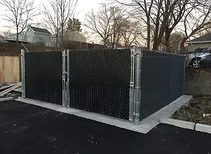 Black chain-link fence with privacy slats encloses a dumpster area on a concrete pad.