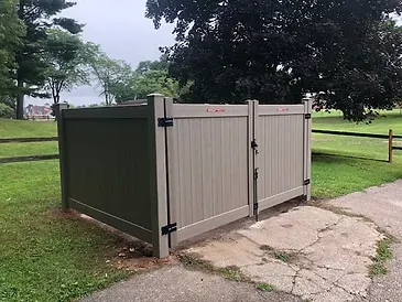 Tan and green dumpster enclosure with closed double doors on a paved area next to grass and trees.