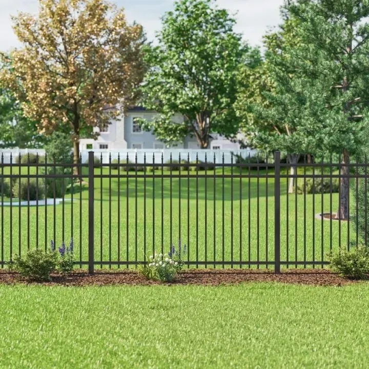 Black metal fence in a green lawn with trees in the background, framing a house on a sunny day.
